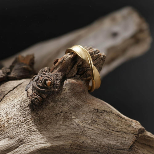 May include: A gold-colored ring with a smooth, curved design. The ring is displayed on a piece of weathered wood, creating a contrast between the polished metal and the natural texture. The ring's shape is simple yet elegant.