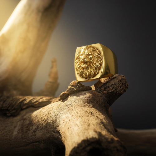 May include: A gold-colored lion ring with a detailed lion's head design. The ring is square-shaped and sits on a piece of wood. The background is dark, with a soft light illuminating the ring.