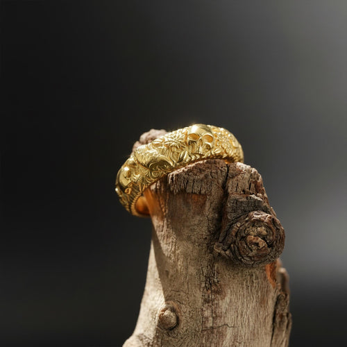 May include: A gold-colored bracelet with intricate carvings of skulls and floral patterns. The bracelet is displayed on a piece of weathered wood against a dark background, highlighting its detailed design and craftsmanship.