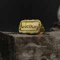 May include: A gold-colored ring with a rectangular face featuring a detailed relief of a religious scene. The ring has an ornate, textured band and is set against a dark background.