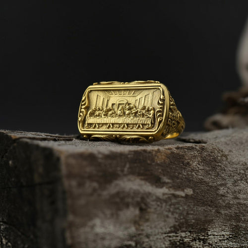 May include: A gold-colored ring with a rectangular face featuring a detailed relief of a religious scene. The ring has an ornate, textured band and is set against a dark background.
