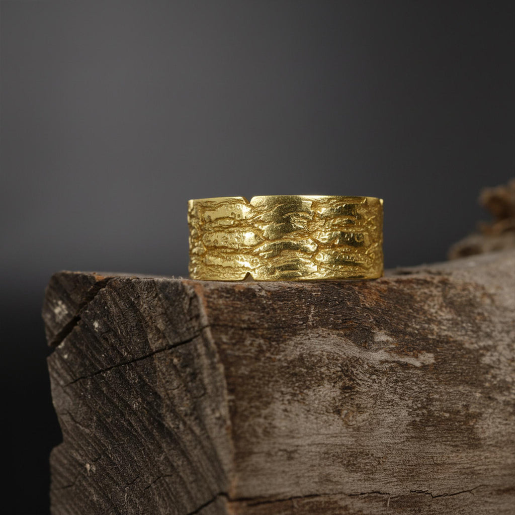 May include: A wide, textured gold ring with a bark-like pattern. The ring is made of a shiny, yellow metal and sits on a piece of weathered wood. The ring's surface has a rough, organic texture.