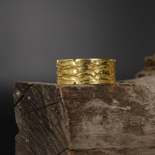 May include: A wide, textured gold ring with a bark-like pattern. The ring is made of a shiny, yellow metal and sits on a piece of weathered wood. The ring's surface has a rough, organic texture.