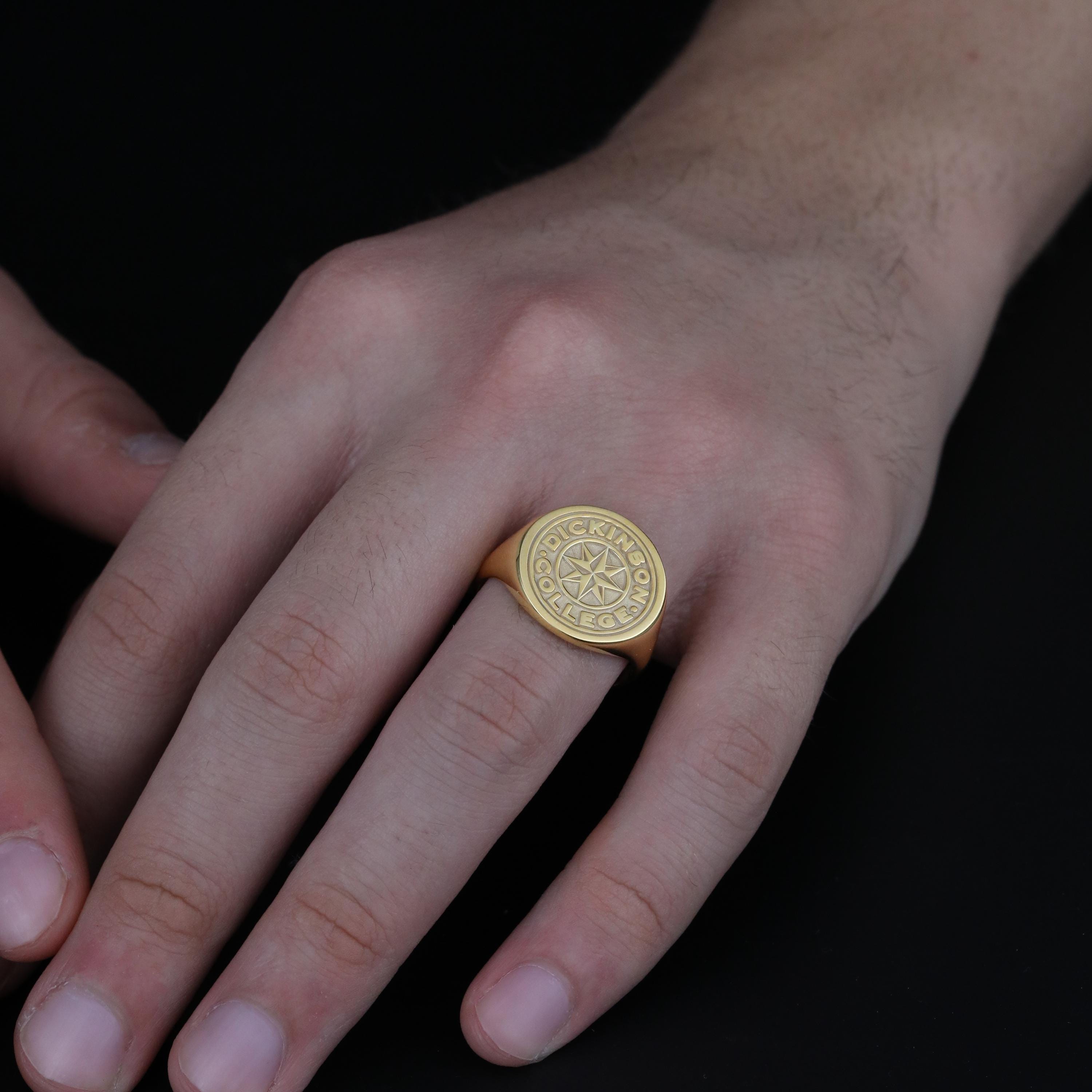 May include: A gold-colored signet ring with the words "DICKINSON COLLEGE" encircling a compass rose design. The ring is worn on a finger, showcasing its polished finish and detailed engraving. The ring is a classic piece of jewelry.