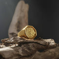 May include: A gold-colored ring with an octagonal face featuring a detailed relief design. The ring is set on a piece of weathered wood against a dark background. The ring's design is reminiscent of a coin.
