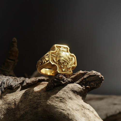 May include: A gold-colored ring with a cross-shaped design. The ring features a detailed relief of a skull wearing a helmet, with ornate scrollwork on the sides. The ring is displayed on a piece of weathered wood against a dark background.