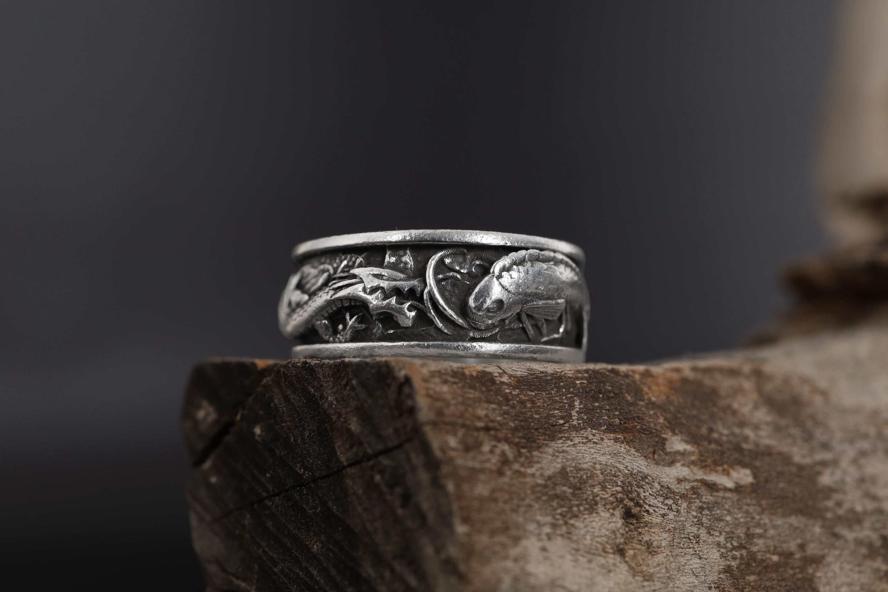 May include: A silver ring with a detailed design of two fish intertwined with floral elements. The ring is on a dark brown wooden surface.